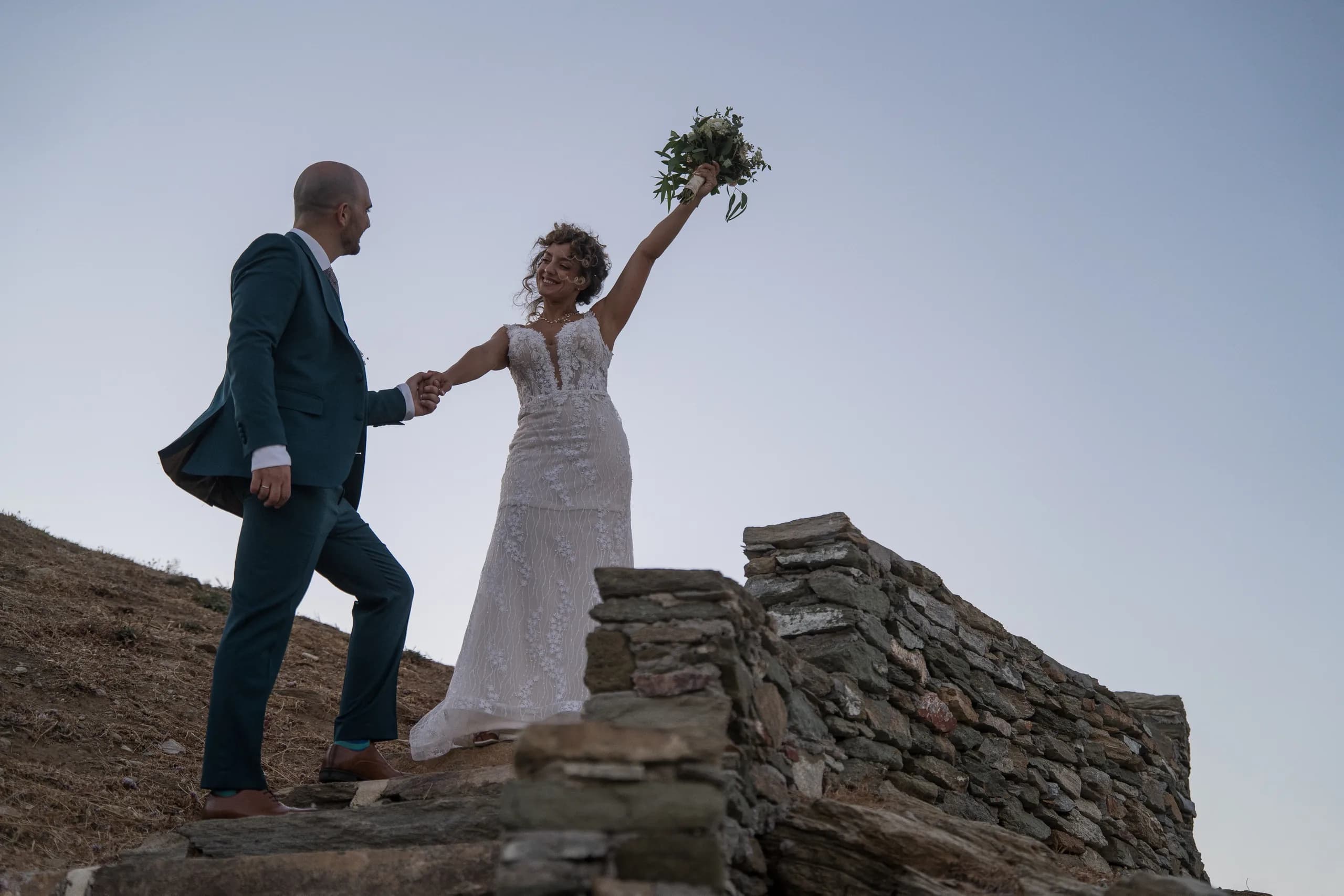 The bride raising her bouquet as the groom leads her up the rocks