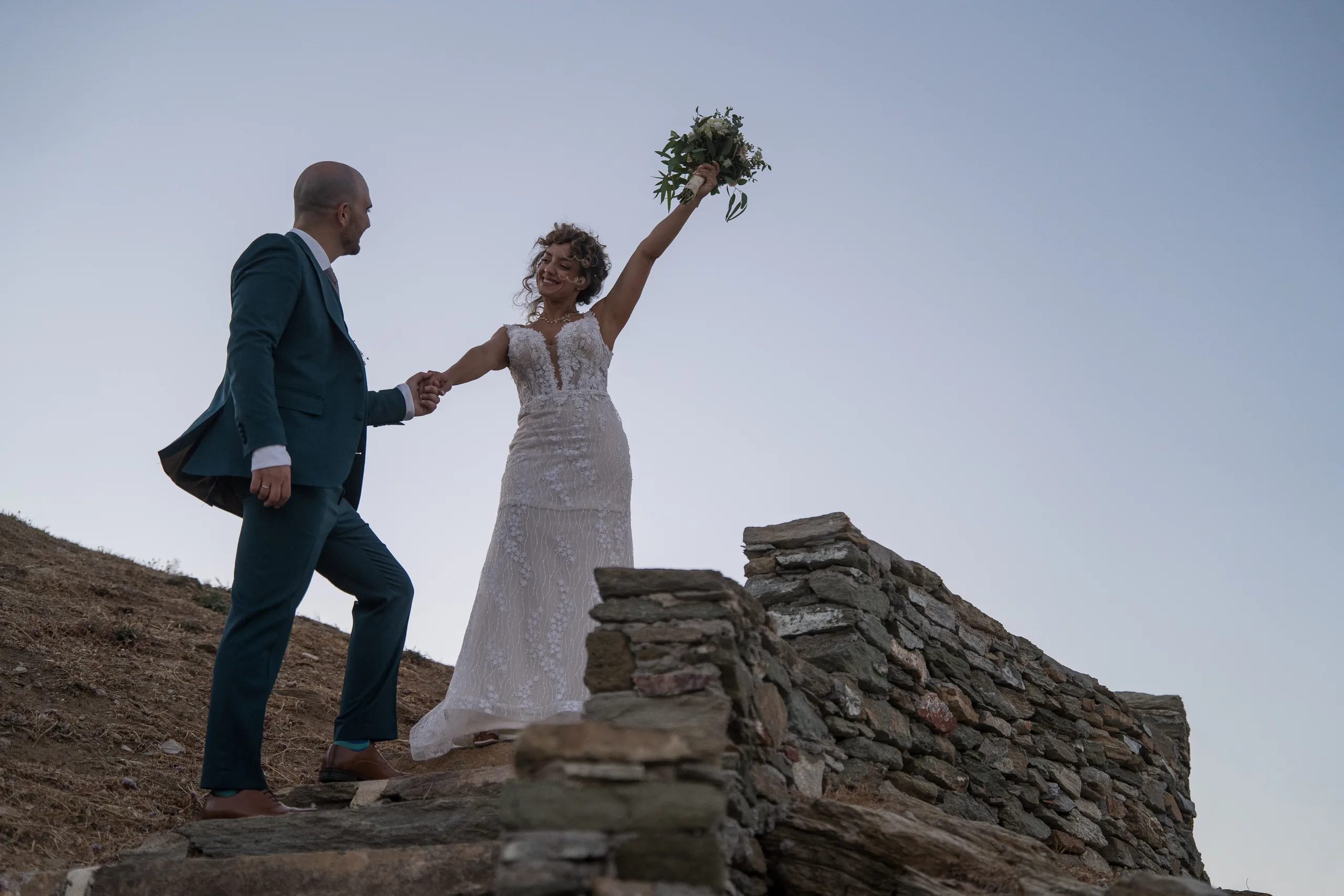 The bride raising her bouquet as the groom leads her up the rocks