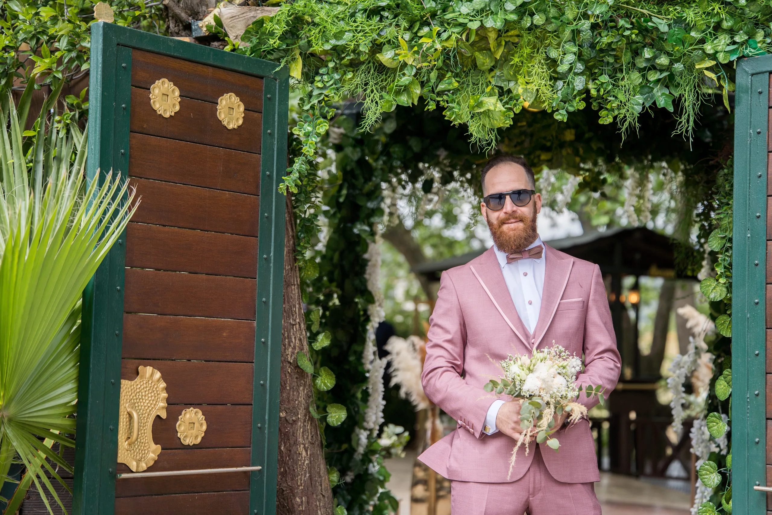 The groom in a pink suit waiting with the bouquet by a wooden door
