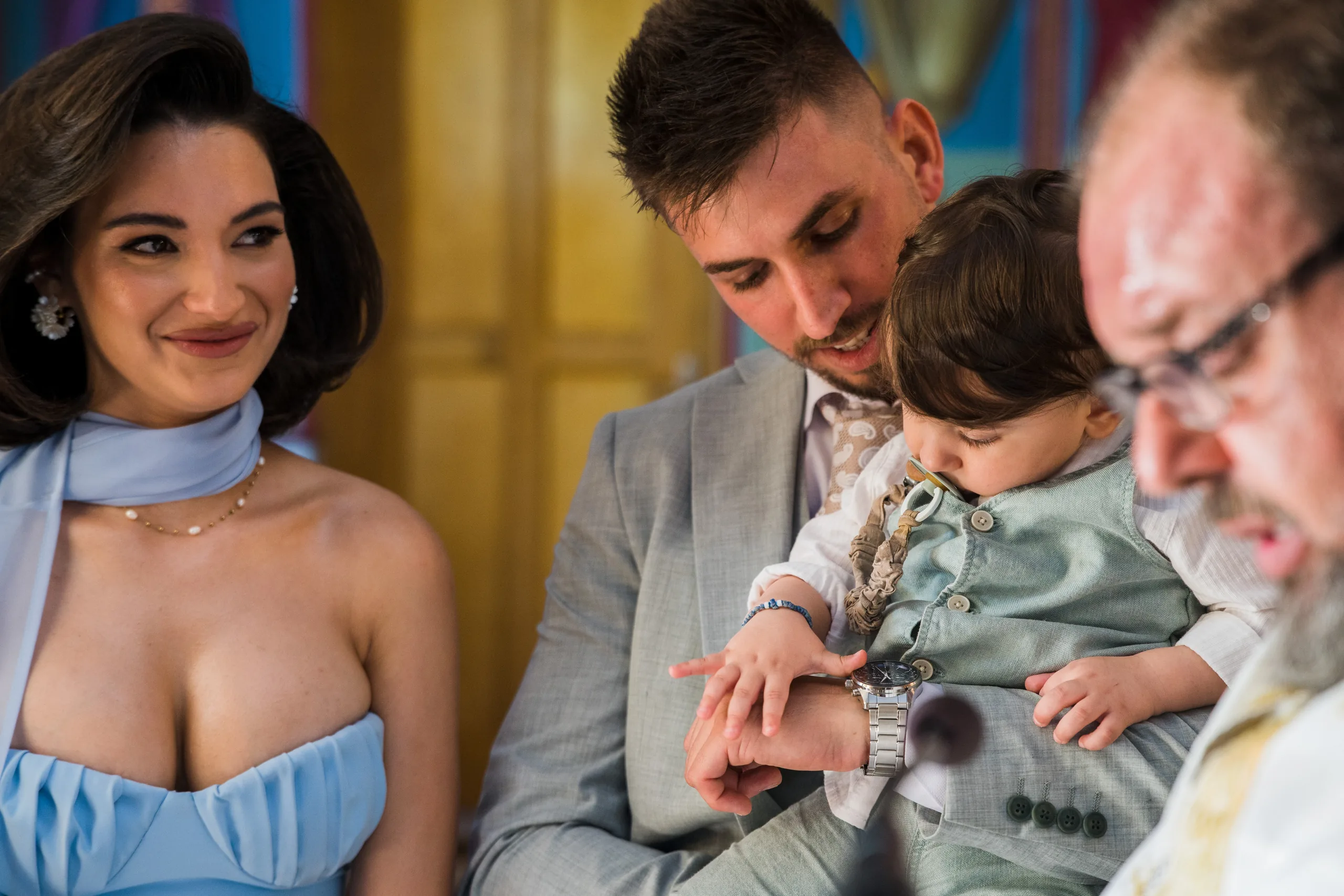 A guest in a blue dress smiles as a man holds the baby beside the priest