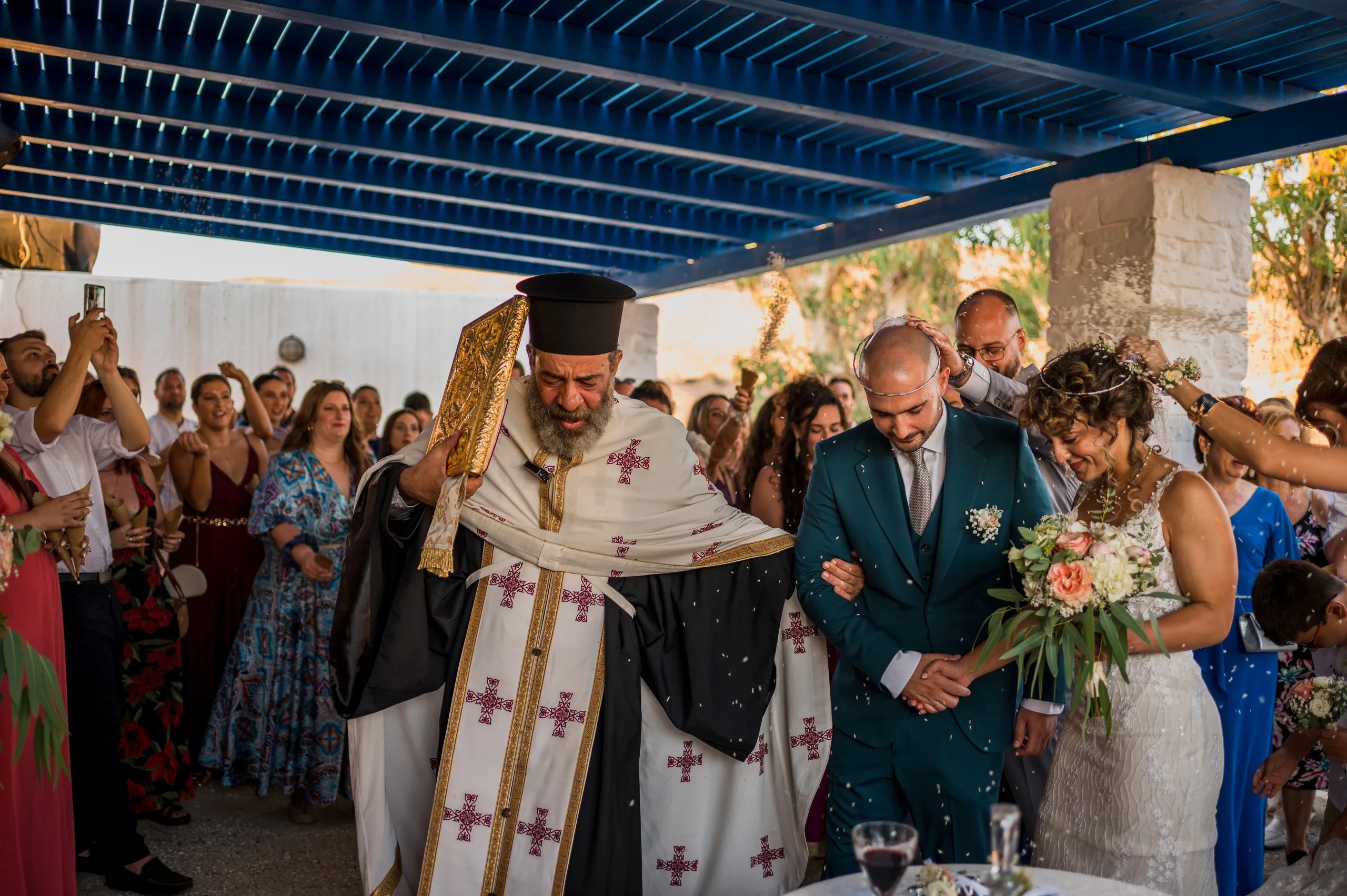 The priest leads the couple around the table under a blue pergola as guests look on