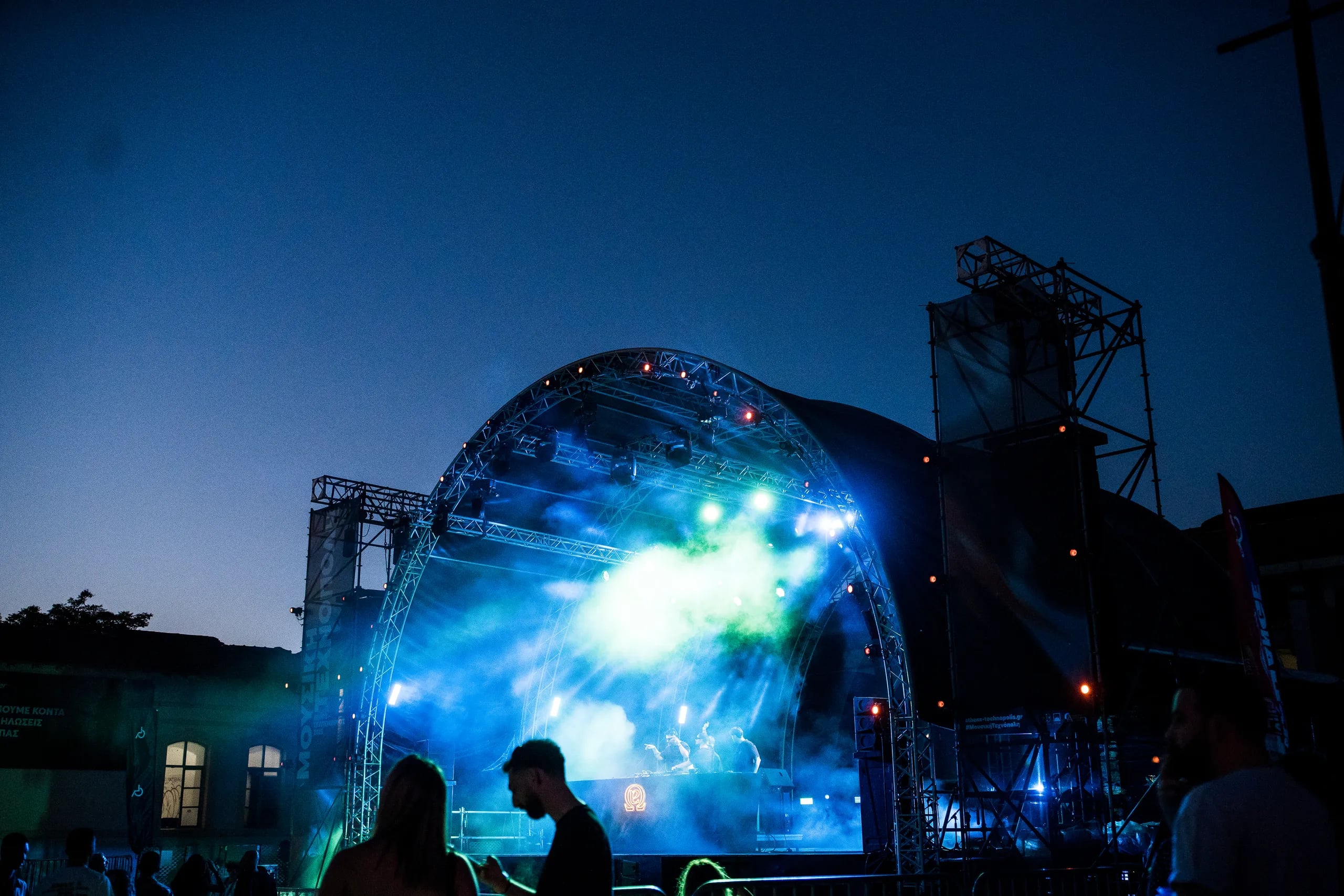 Outdoor concert stage at dusk, lit in blue and green, with the crowd in silhouette
